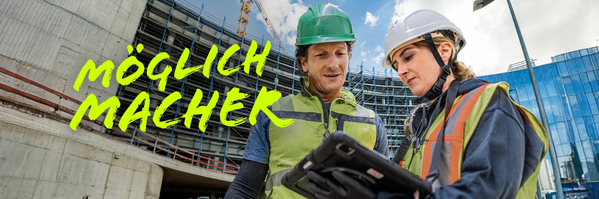 Ein Mann und eine Frau in Schutzkleidung sprechen über ein Projekt auf dem Tablet, im Hintergrund ist eine große Baustelle.